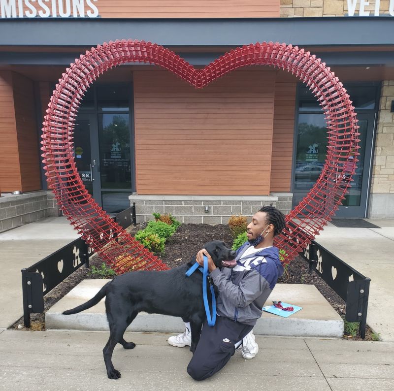 man crouched down with his large black dog in front of the KC Pet Project Heart Arch.