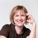 Heather Lewis in a brown shirt with blonde hair. She has one hand up with her fingertips touching her face.