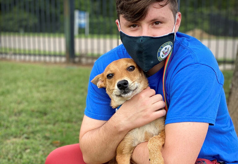young man wearing mask hugs medium sized tan dog