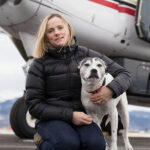 Kara Pollard with a dog in front of an airplane. Kara is wearing a black puffer jacket and jeans.